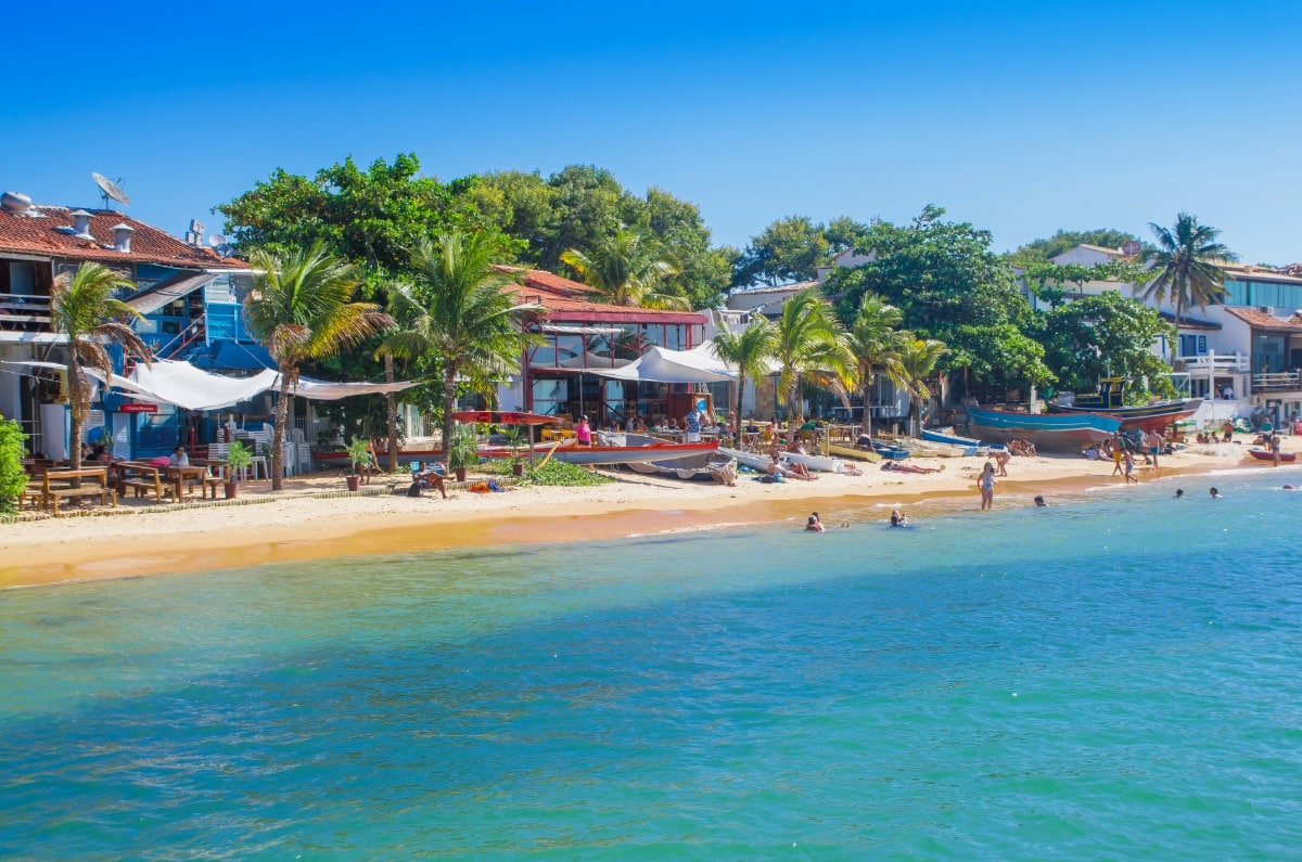 Vista de la costa de Búzios, Brasil, con pequeñas embarcaciones, arena dorada, aguas transparentes y restaurantes frente al mar.