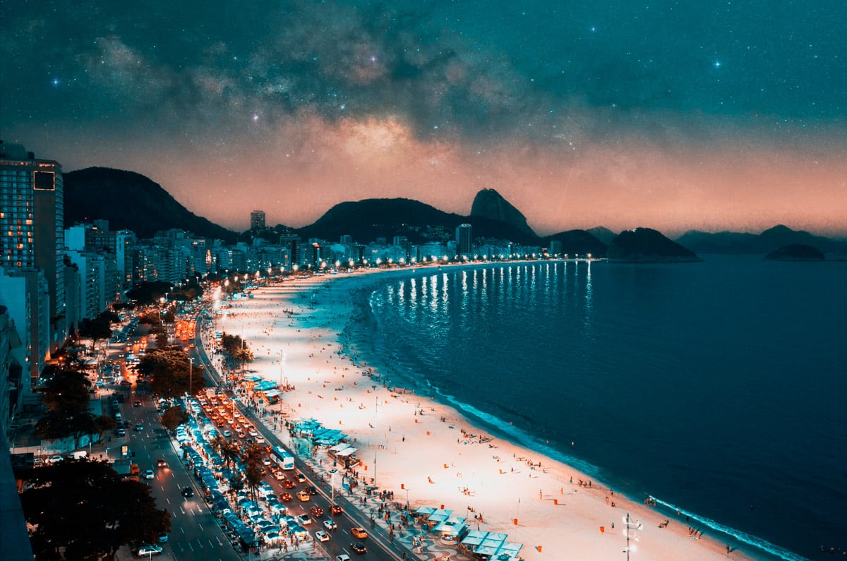 Vista nocturna de la playa de Copacabana en Río de Janeiro iluminada bajo un cielo estrellado con la Vía Láctea y el Pan de Azúcar al fondo.