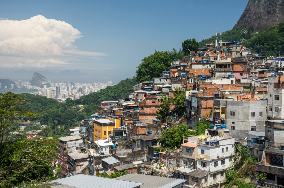Vista panorámica de la Favela Rocinha en Río de Janeiro, mostrando su arquitectura orgánica sobre la colina con el skyline de la ciudad y el Pan de Azúcar al fondo.