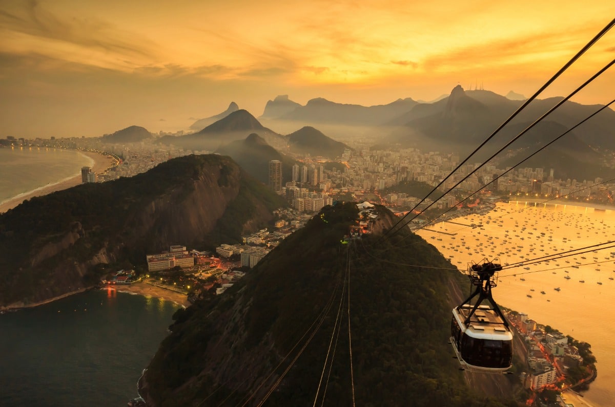 Teleférico del Pan de Azúcar sobrevolando la Bahía de Guanabara en Río de Janeiro al atardecer, con vistas a la playa de Botafogo y las montañas.