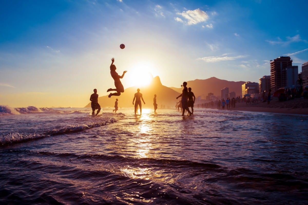 Jóvenes brasileños jugando al fútbol playa al atardecer en Ipanema, con la silueta del Morro Dois Irmãos de fondo en Río de Janeiro.