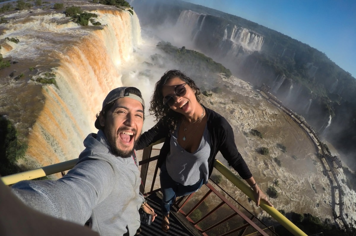 Pareja sonriente haciéndose un selfie en la pasarela sobre las aguas de las Cataratas del Iguazú durante su luna de miel en Brasil.