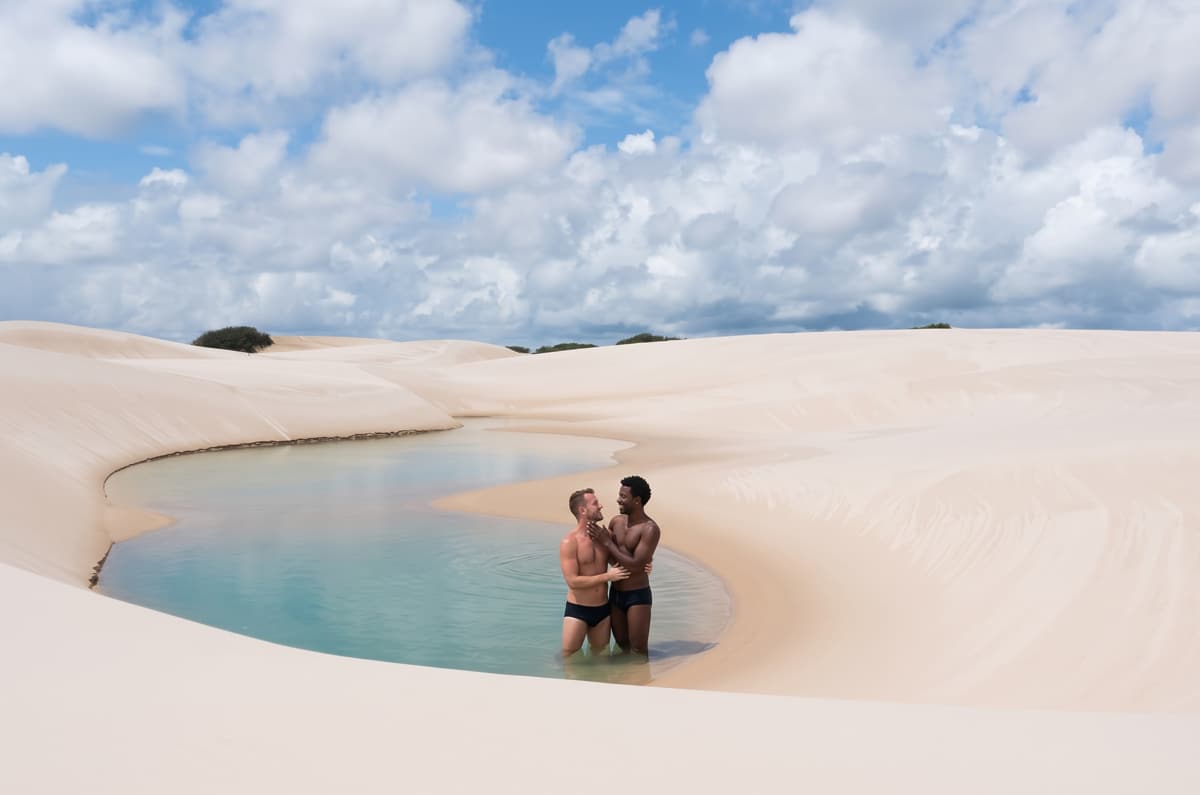 Pareja de hombres abrazados dentro de una laguna cristalina rodeada de dunas blancas durante su luna de miel en Lençóis Maranhenses, Brasil.