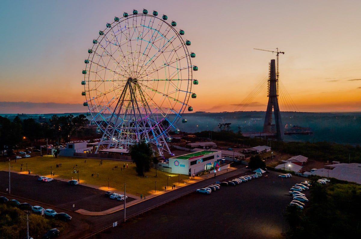 Noria panorámica Yupi Star en Foz do Iguaçu iluminada al atardecer, junto al puente de integración fronteriza entre Brasil y Paraguay.