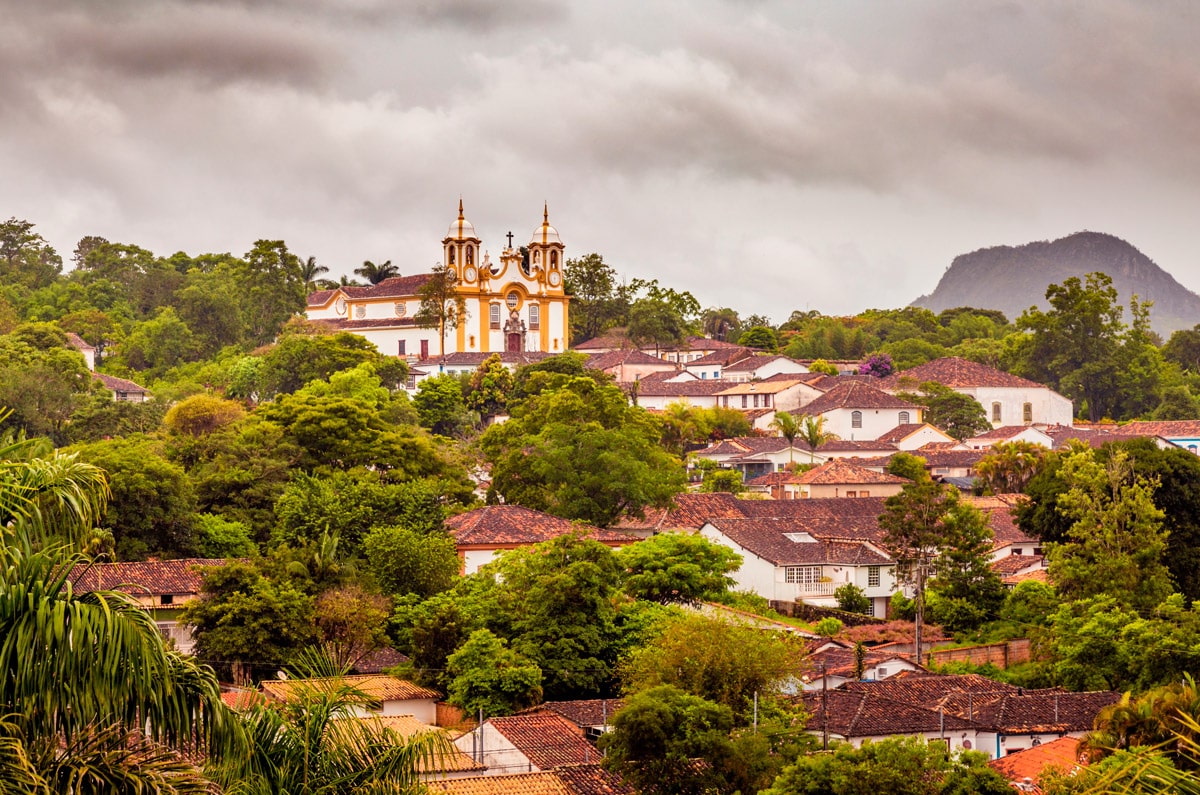 Vista panorámica de la ciudad colonial de Tiradentes en Minas Gerais, Brasil, con su iglesia barroca rodeada de vegetación bajo un cielo nublado.