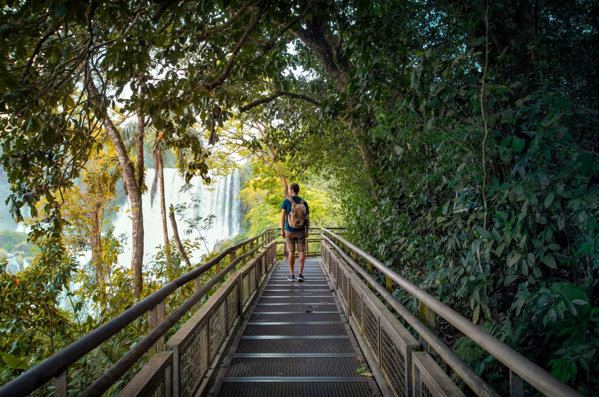 Viajero con mochila recorriendo las pasarelas de las Cataratas del Iguazú en el lado de Brasil, con las caídas de agua al fondo rodeadas de selva tropical.